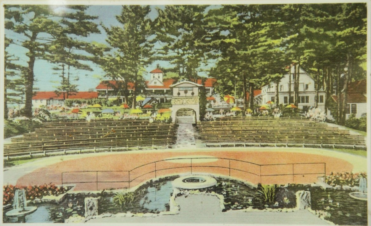 An old fashioned color postcard shows an outdoor amphitheater in front of a white and red-trimmed resort.
